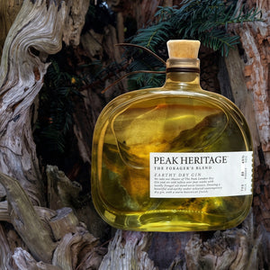 A bottle of Peak Heritage Captive Dry Gin with a cork stopper, filled with golden amber liquid, set against a rustic background of weathered tree bark and pine needles.