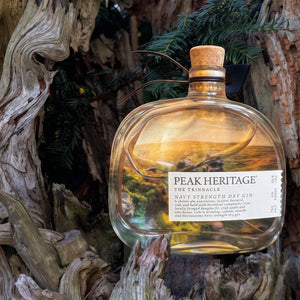A bottle of Peak Heritage Navy Strength Flat Gin with a scenic landscape label, set against a rustic background of weathered tree bark and pine branches.